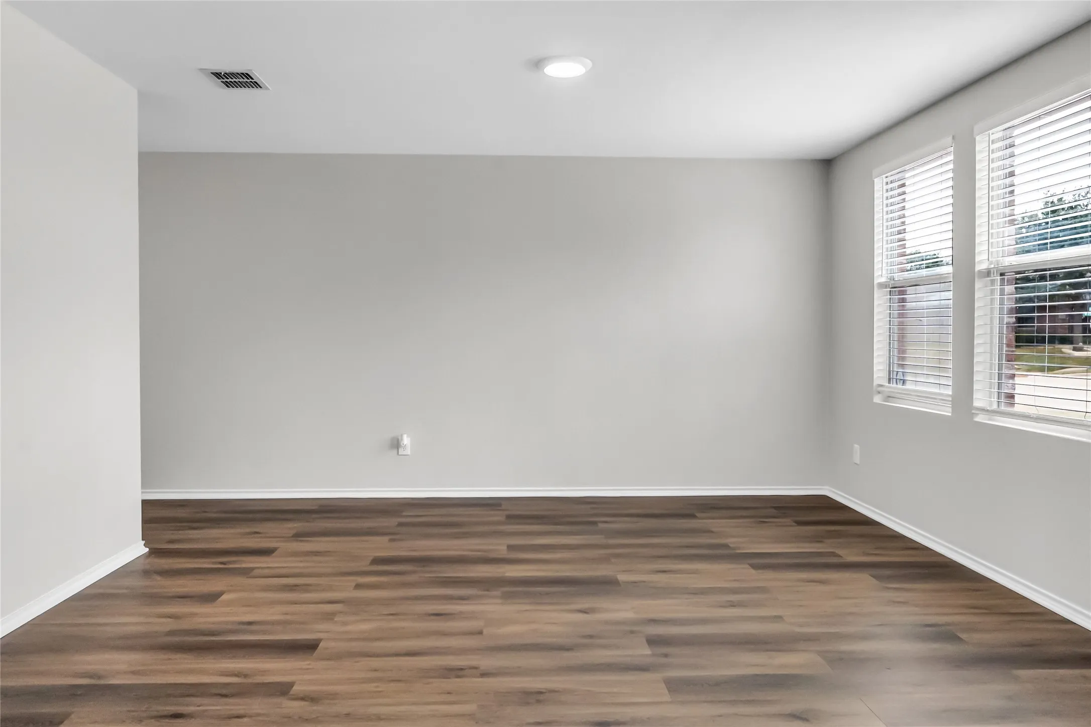 Expansive room featuring wood-finish flooring, light gray wall tones, two windows with blinds, and recessed lighting