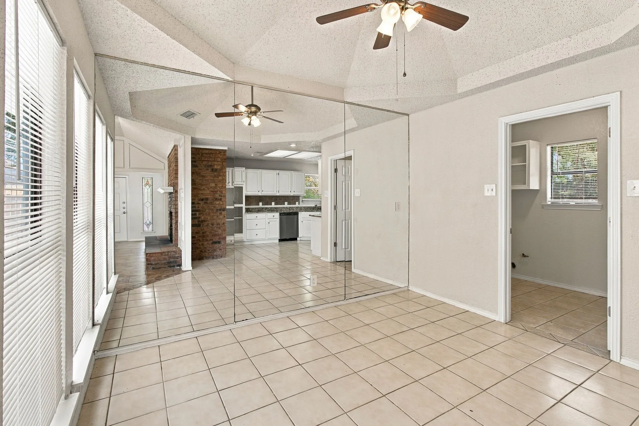 Vaulted ceiling with ceiling fan, tile flooring, mirrored wall, brick fireplace surround, and white kitchen cabinetry