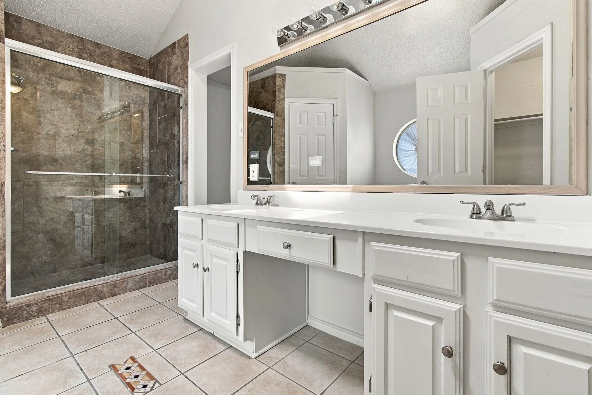 Dual vanity with integrated sinks, framed mirror, glass-enclosed shower with tiled surround, and tiled flooring