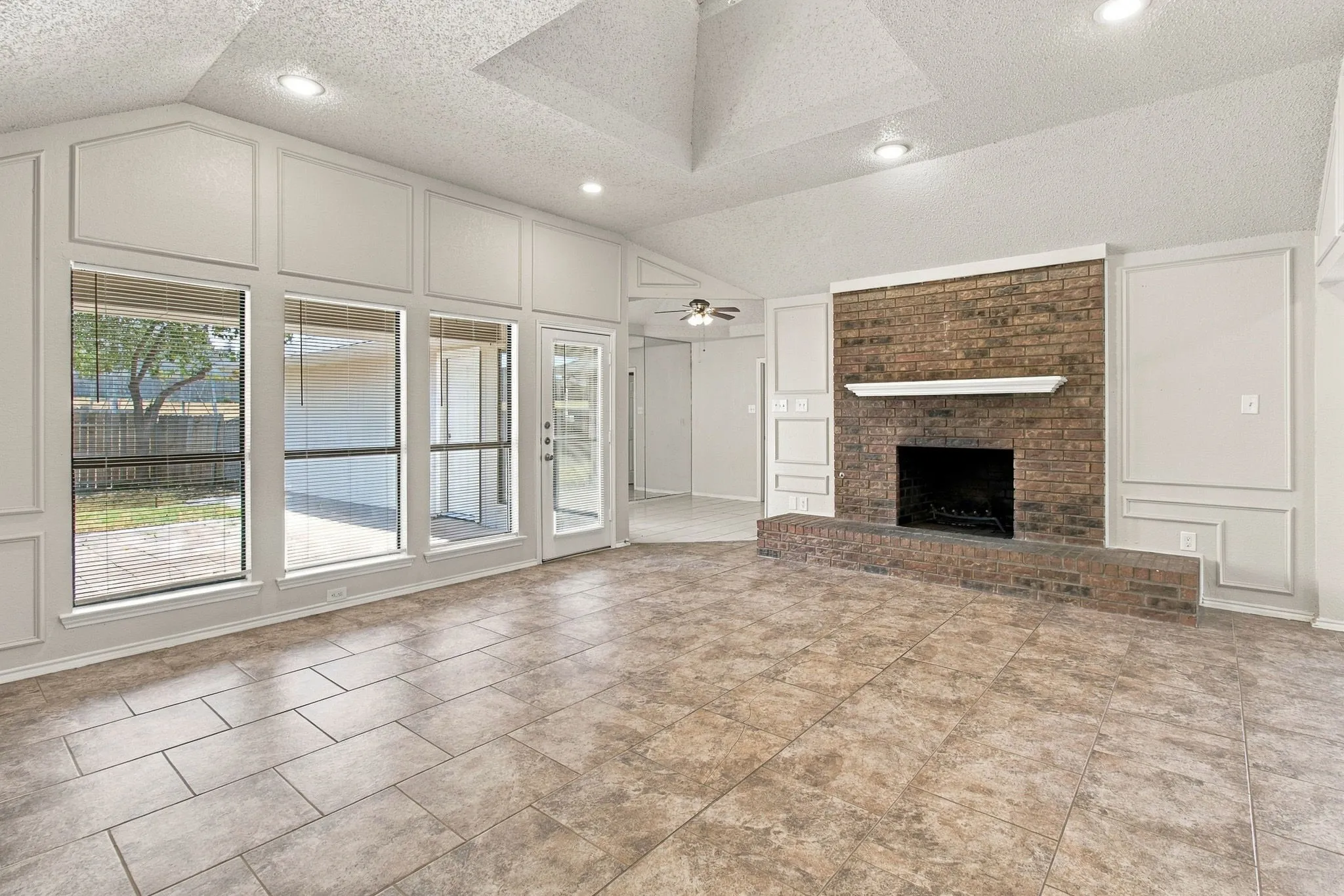 Vaulted ceiling with recessed lighting, brick-faced fireplace with hearth and mantel, wall paneling, large windows, and tile flooring