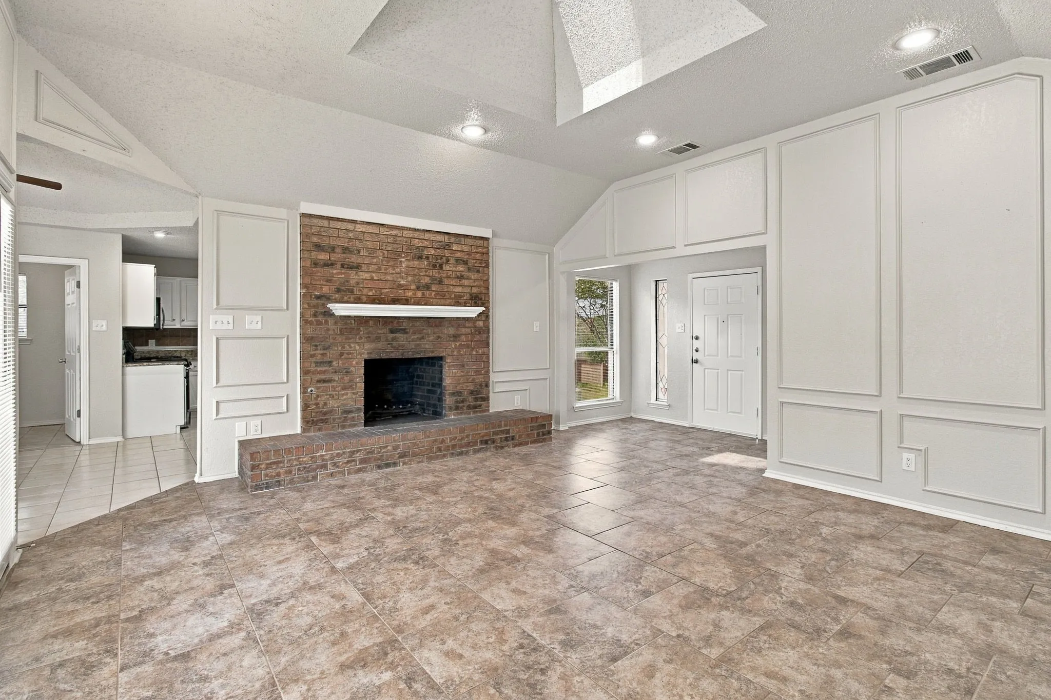 Vaulted ceiling with skylight, brick fireplace with mantel, decorative wall paneling, tile flooring, and recessed lighting