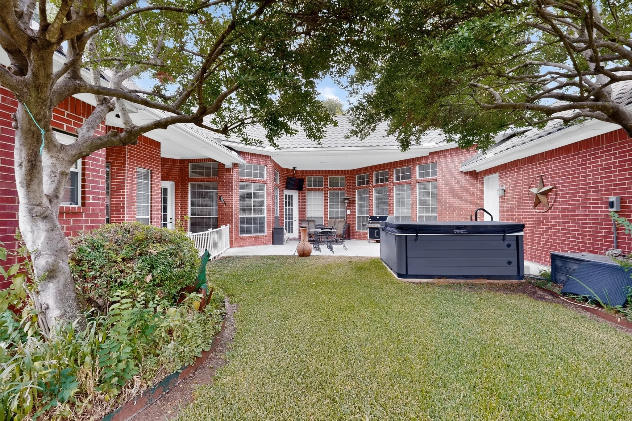 Expansive backyard featuring a manicured lawn, red brick exterior, mature trees, and a built-in spa
