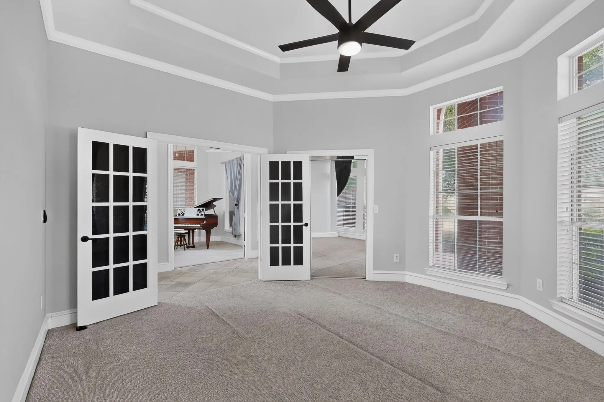 Tray ceiling with crown molding, double French doors, large windows with blinds, and a ceiling fan with integrated lighting