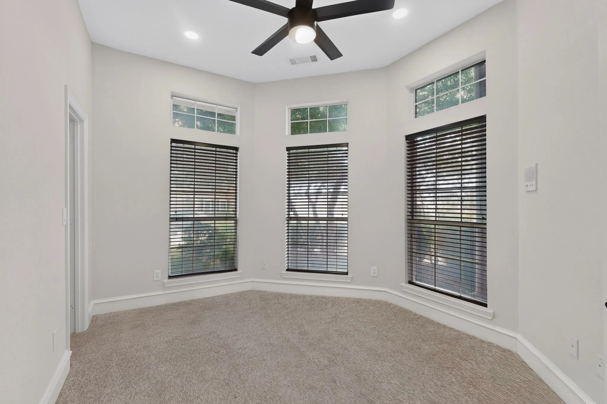Light gray walls with white trim, gray carpet flooring, a black ceiling fan, and multiple windows with horizontal blinds