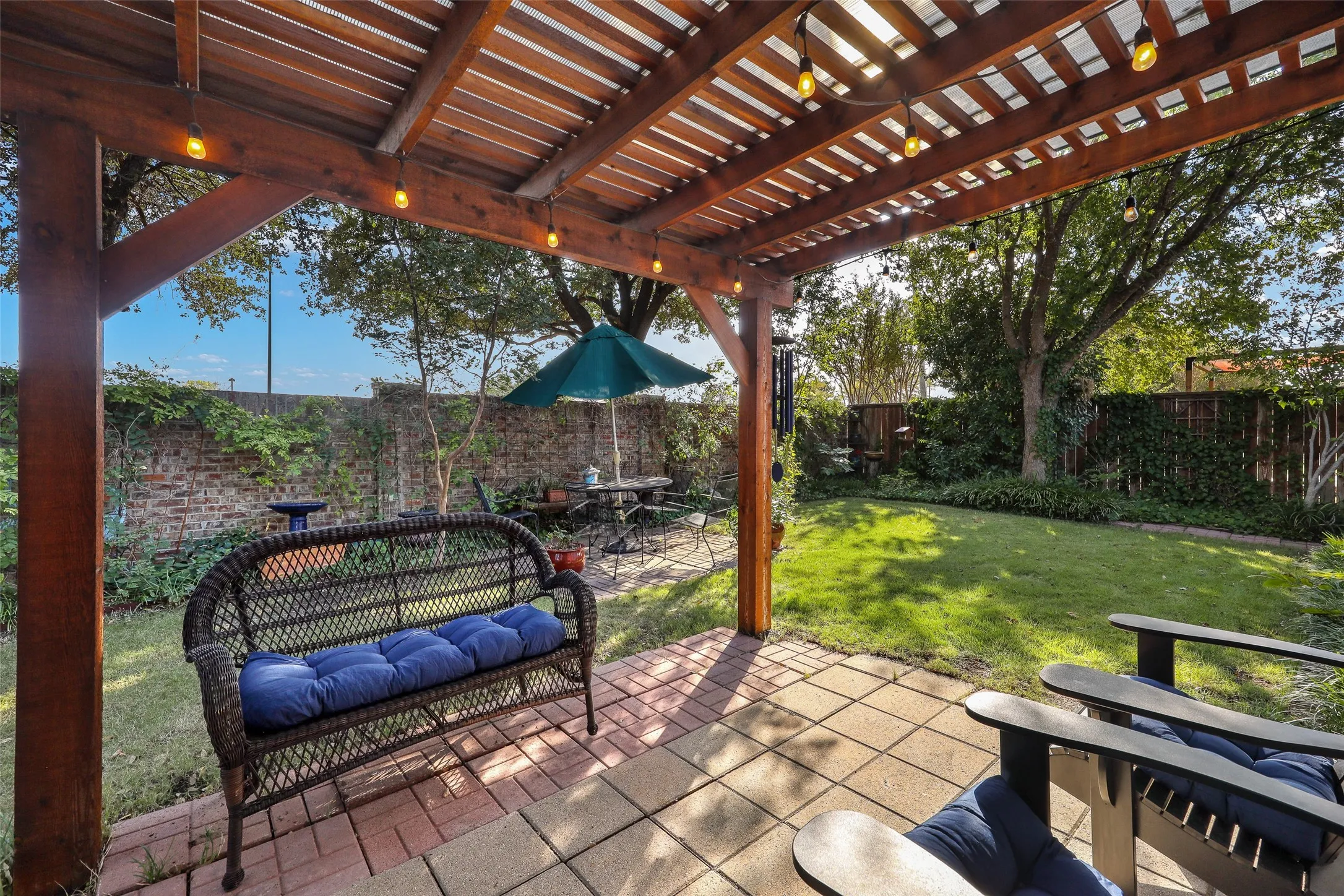 Wood pergola with string lights, brick paver patio, brick perimeter wall, and a fenced yard with mature landscaping