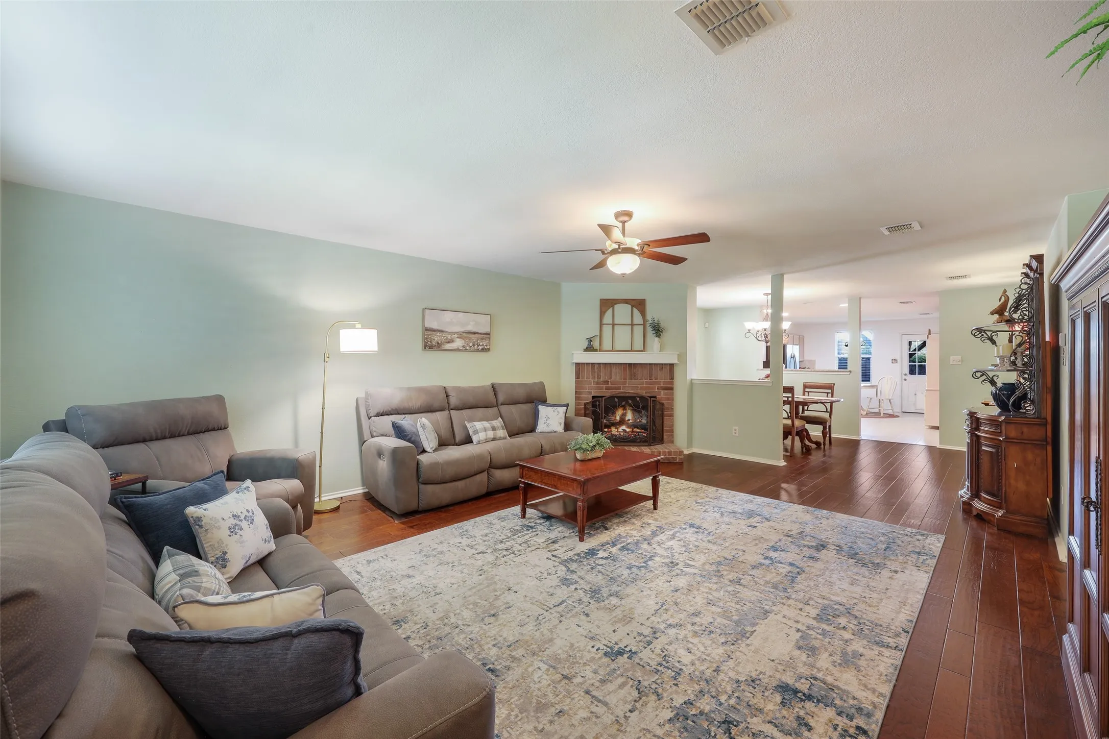 Spacious living room with hardwood flooring, ceiling fan, and fireplace with brick surround