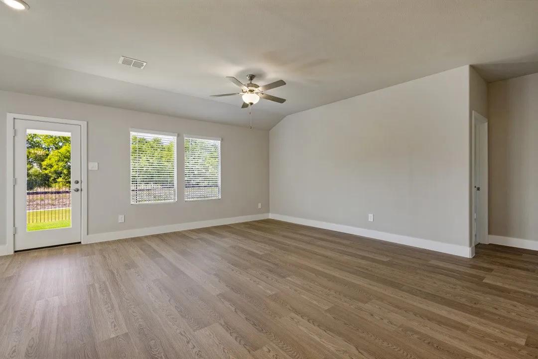 Spacious room featuring wood-finish flooring, multiple windows with blinds, a ceiling fan with integrated lighting, a white door providing outdoor access, and neutral wall tones