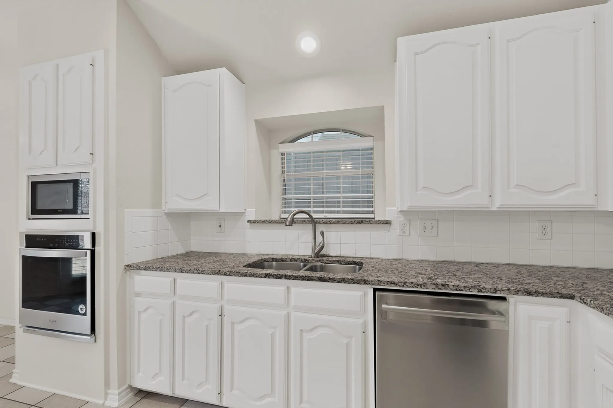 White cabinetry with arch details, stainless steel double basin sink, stainless steel dishwasher, speckled countertops, and white tile backsplash