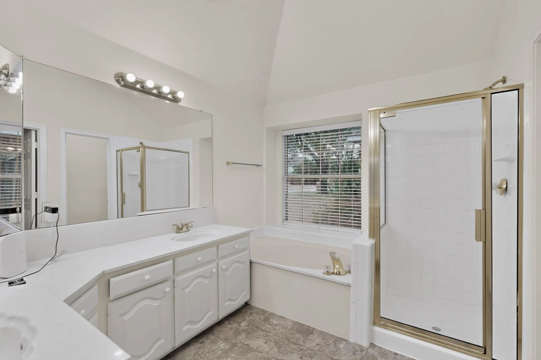 Vaulted ceiling with overhead lighting, large vanity mirror, white cabinetry with integrated sinks, and a walk-in shower with a gold-tone frame