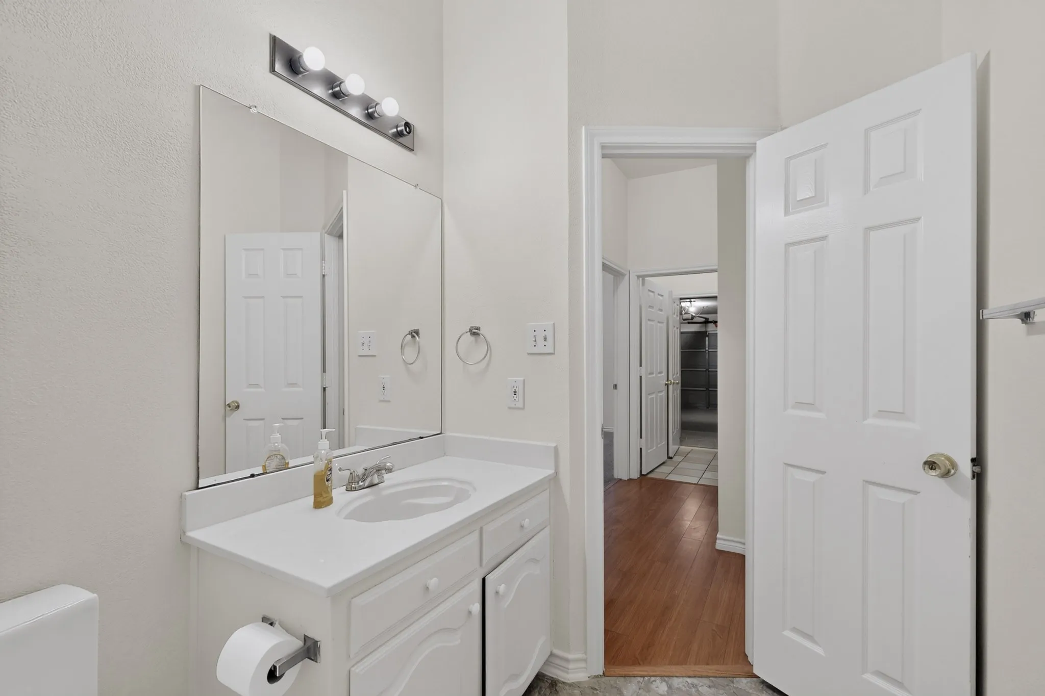 White vanity with an integrated sink and chrome faucet, illuminated by a four-bulb vanity light, with wood-finish flooring extending from the doorway