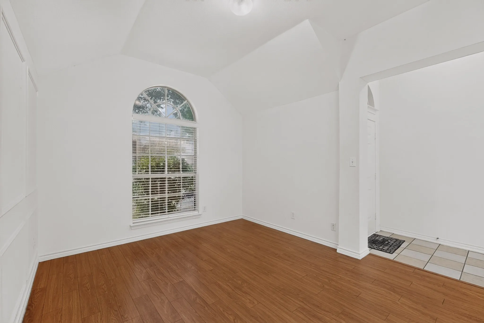 Vaulted ceiling, arched window with blinds, wood-finish flooring, white paneled wall, tiled entry