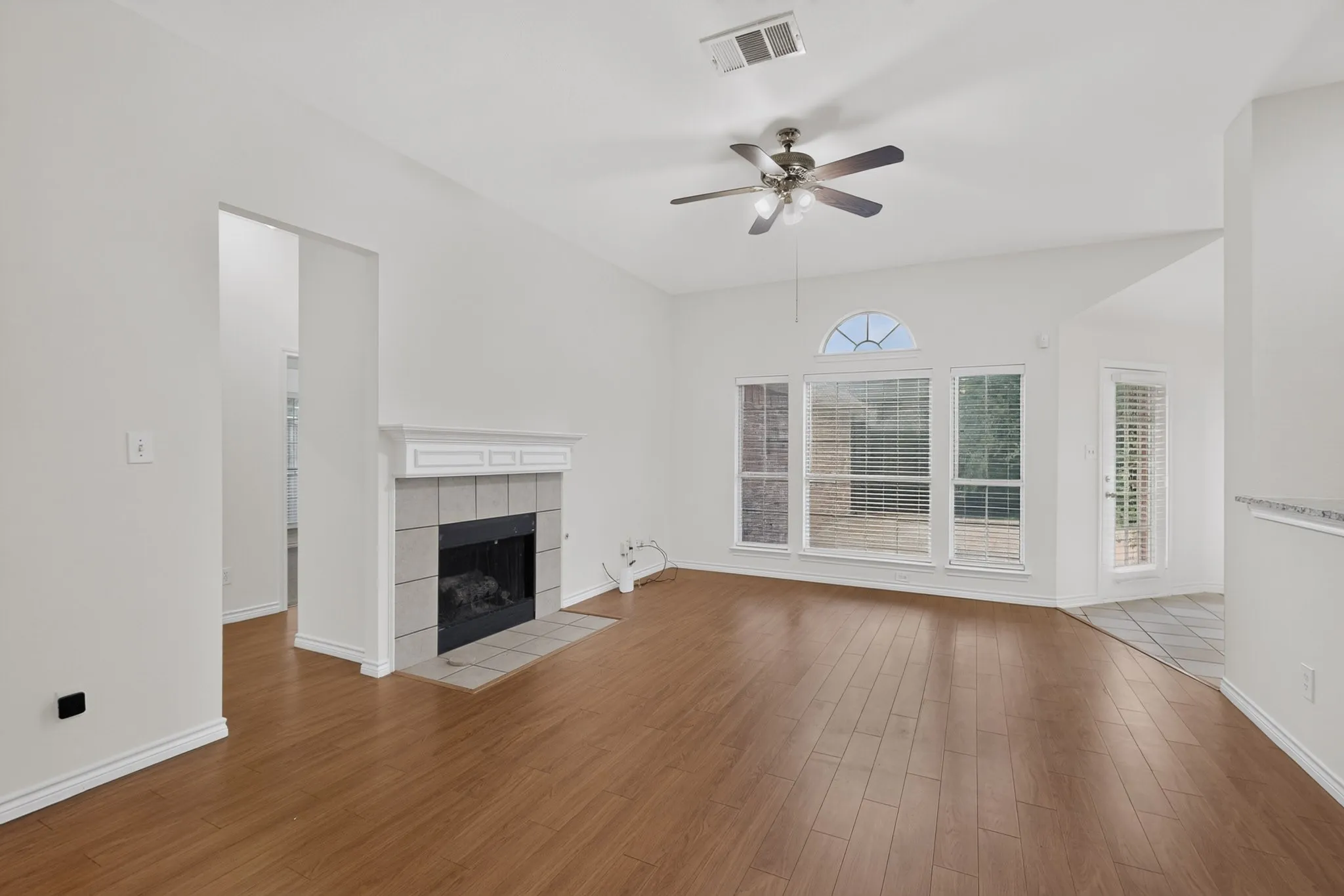 High vaulted ceiling with a ceiling fan, wood-finish flooring, tiled fireplace with a mantel, large windows with an arched transom, and a glass-paneled exterior door