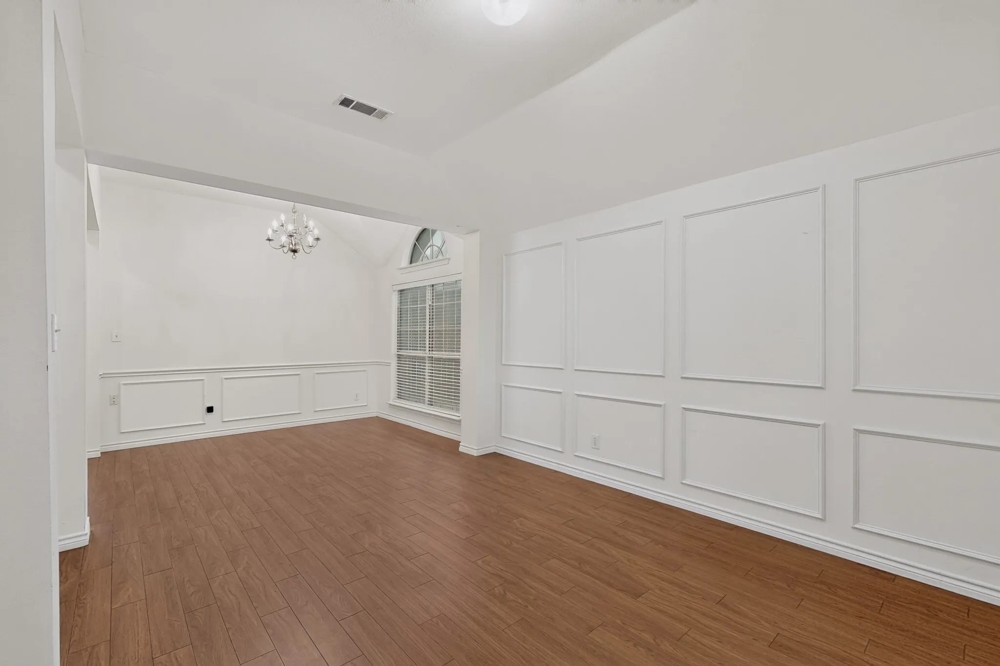 Wood-finish flooring, accent wall paneling, vaulted ceiling, arched window, and a built-in chandelier