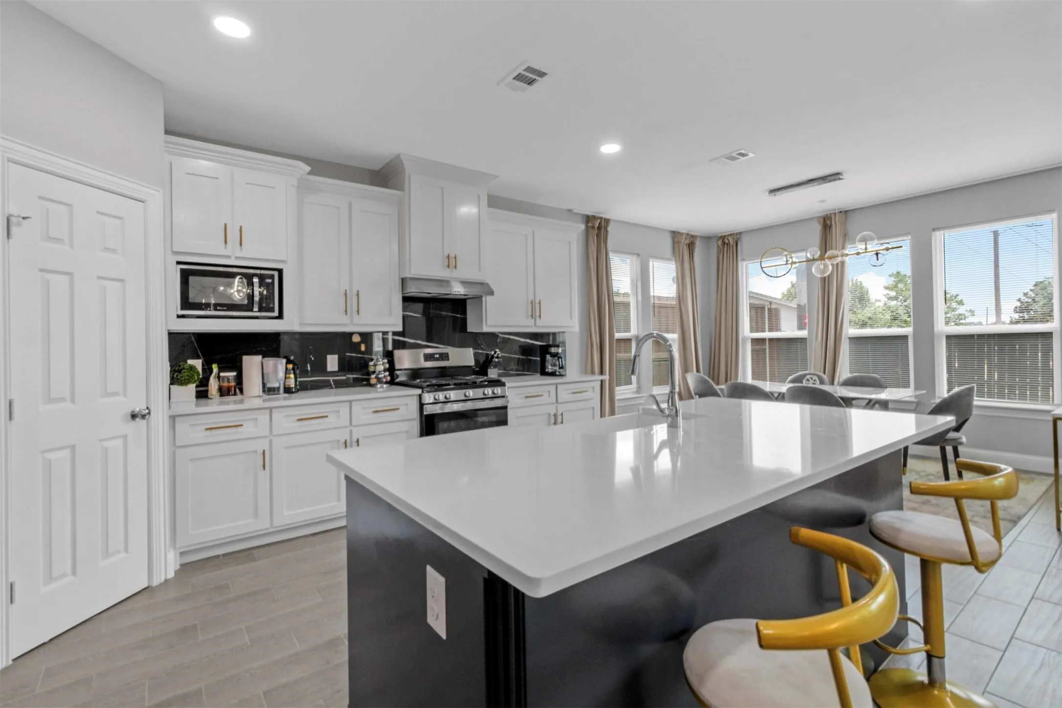 White cabinetry with gold-tone hardware, stainless steel appliances, dark stone backsplash, central island with an integrated sink, and wood-finish flooring