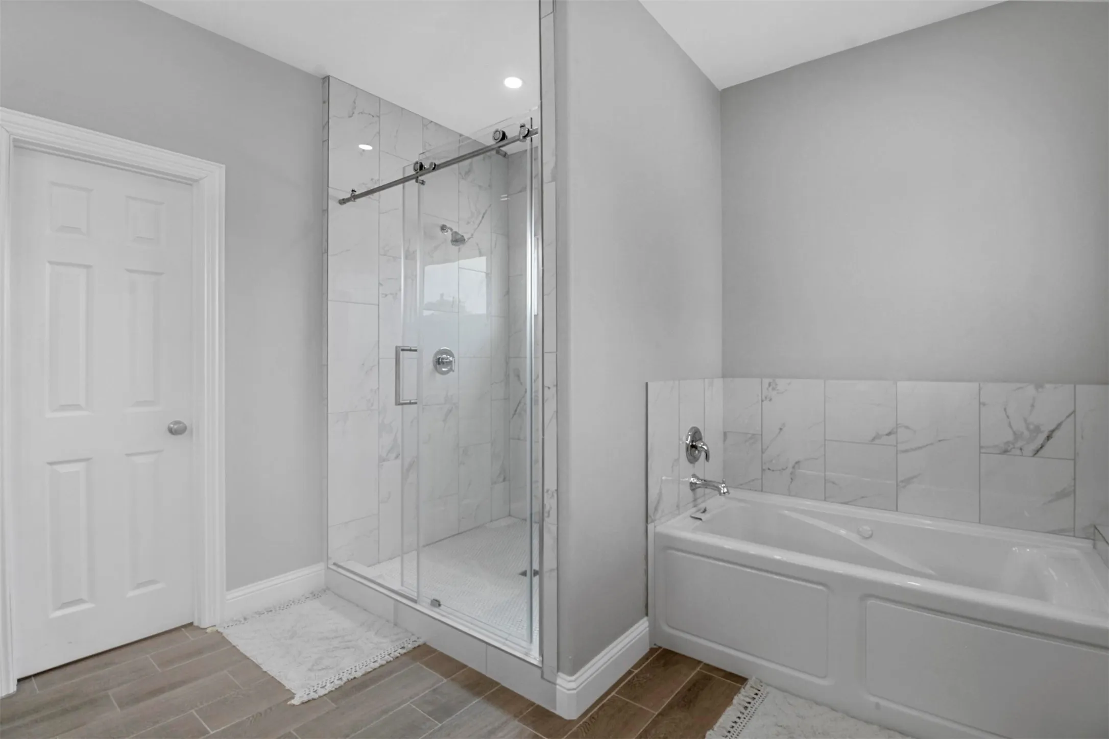 Walk-in shower with clear glass enclosure, a built-in tub with tile surround, and wood-finish flooring