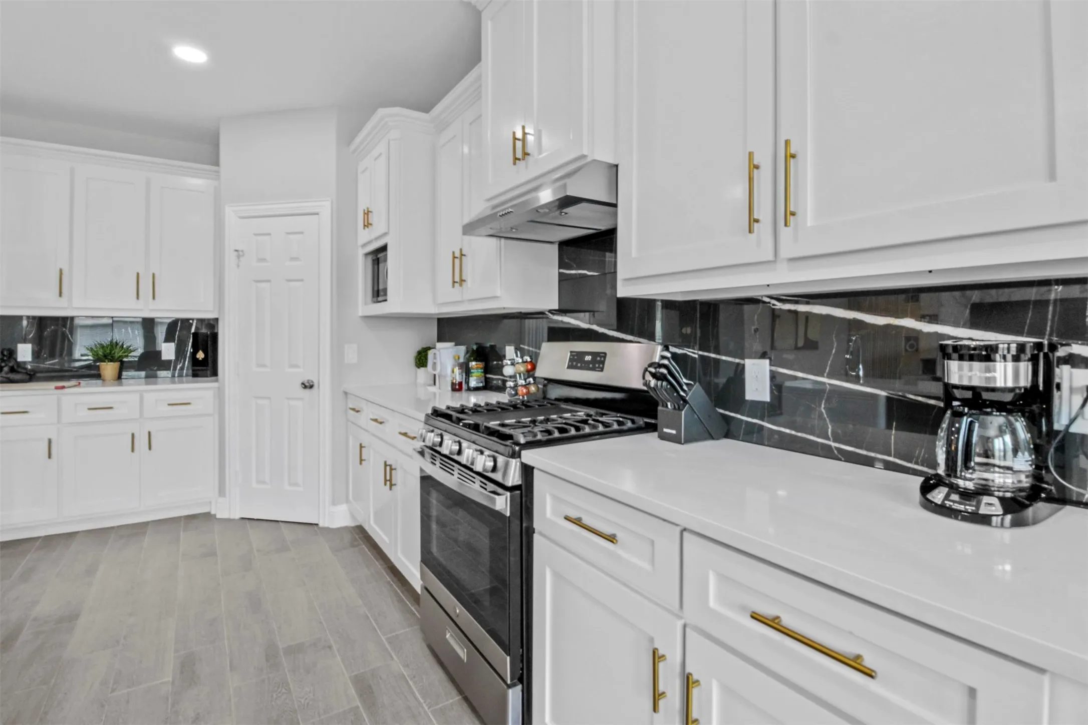 White cabinetry, stainless steel gas range, black backsplash with white veining, light-toned countertops, and wood-finish flooring