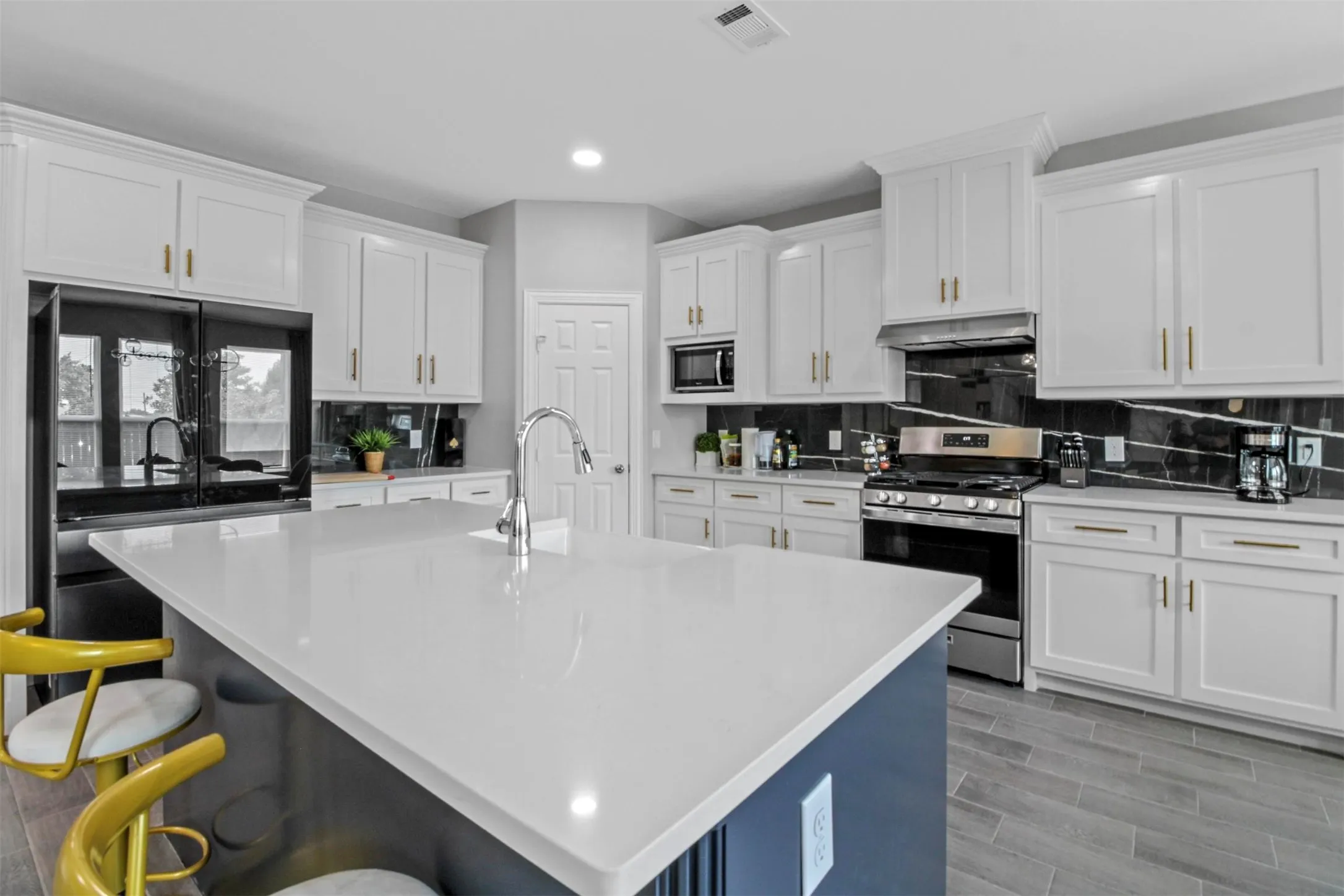 Spacious kitchen featuring an expansive island with a light-toned countertop, white cabinetry with gold-toned hardware, a stainless steel range with a coordinating hood, and a dark-toned backsplash