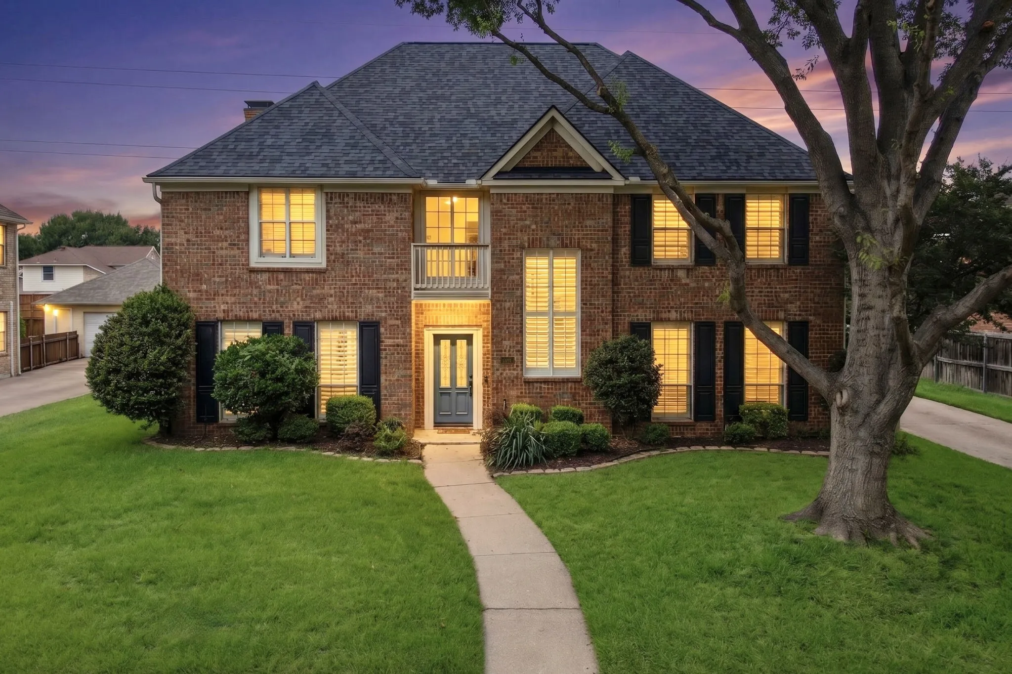 Brick facade residence featuring a dark shingled roof, black louvered shutters, an upper-level balcony, and a manicured lawn with mature landscaping