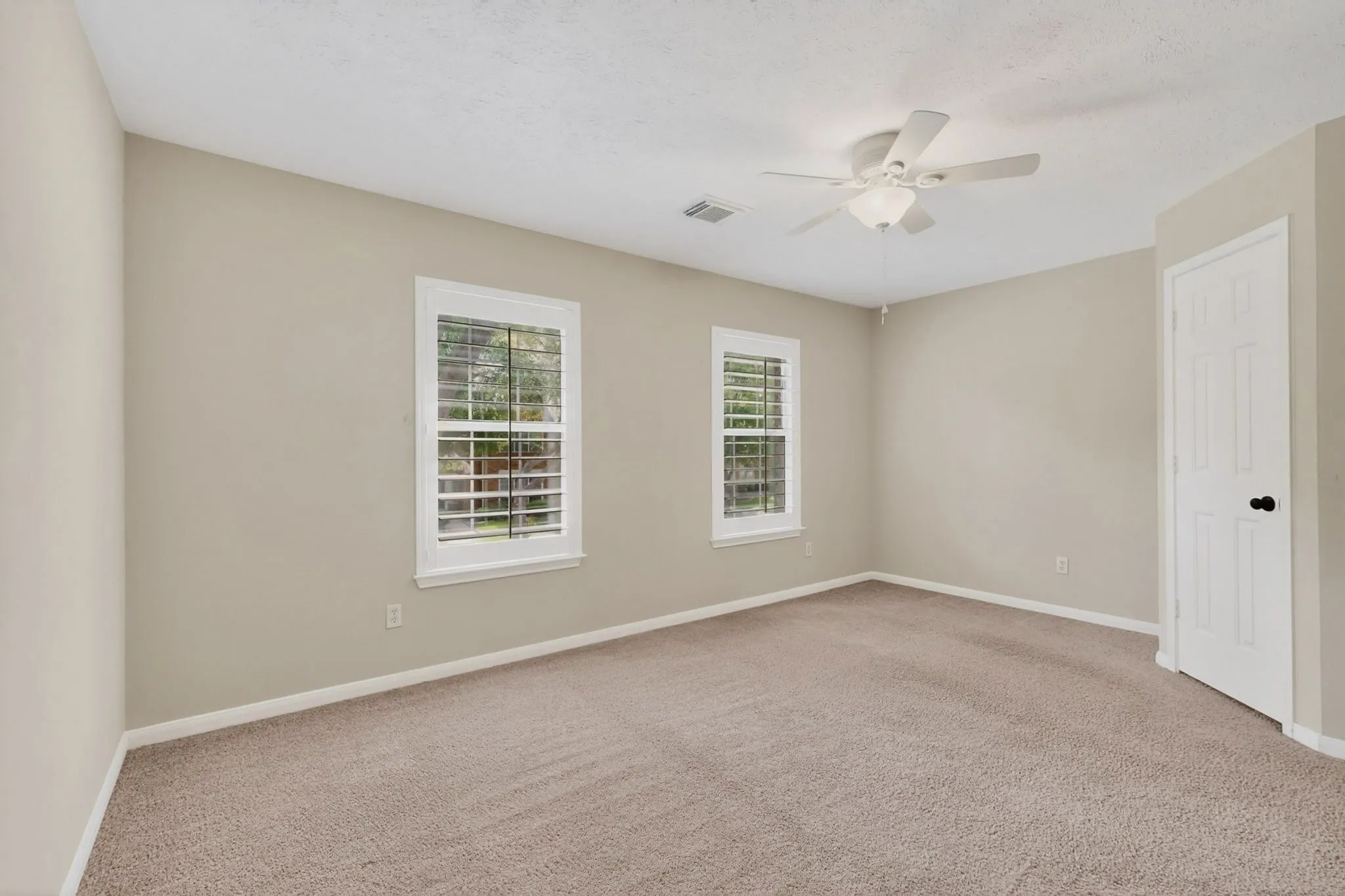 Neutral wall tones, carpeted flooring, two windows with white plantation shutters, white ceiling fan with integrated lighting, and a white paneled door with black hardware