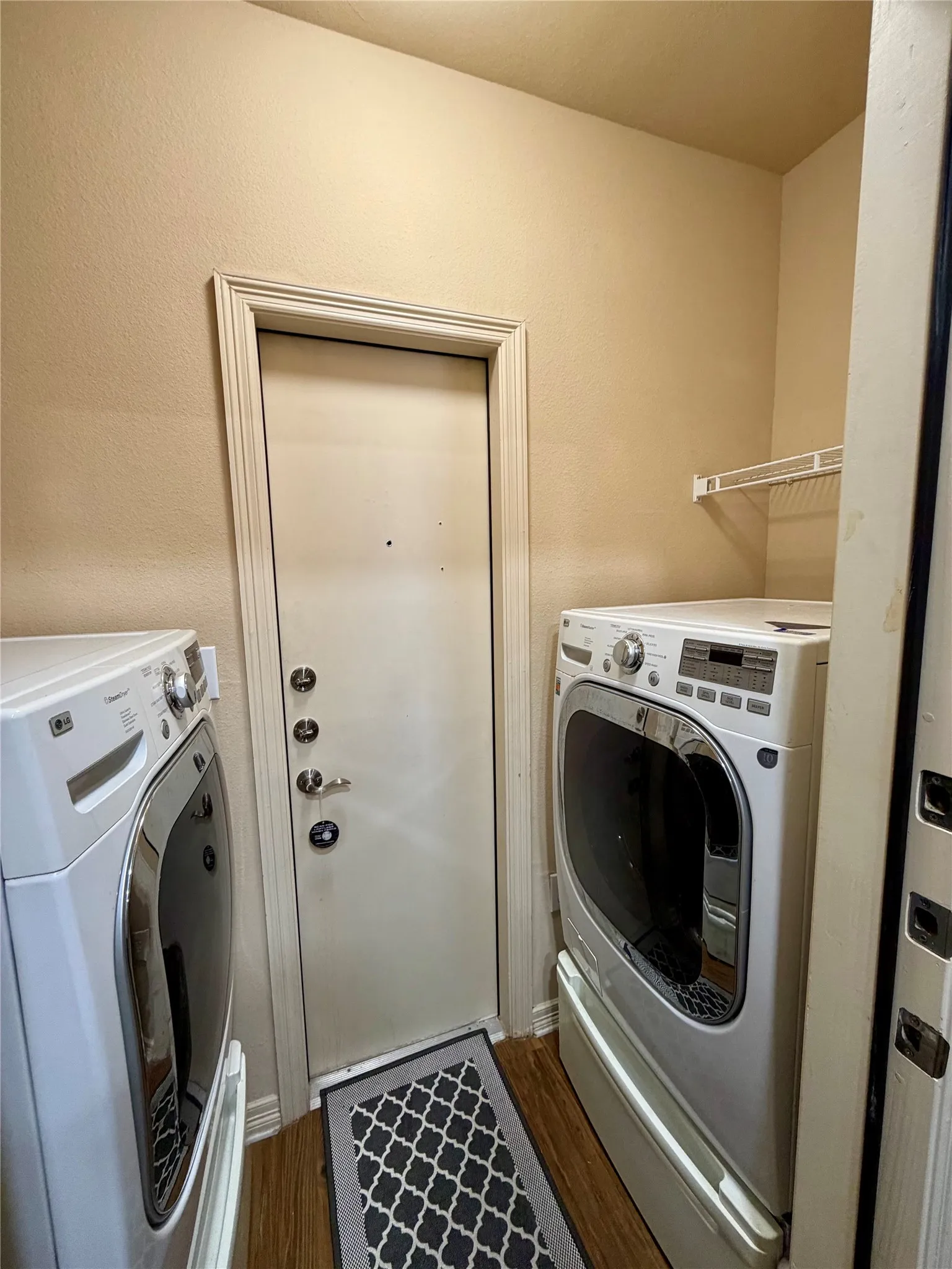 Laundry area featuring wood-finish flooring, a white metal wire shelf, and a white door with multiple locks