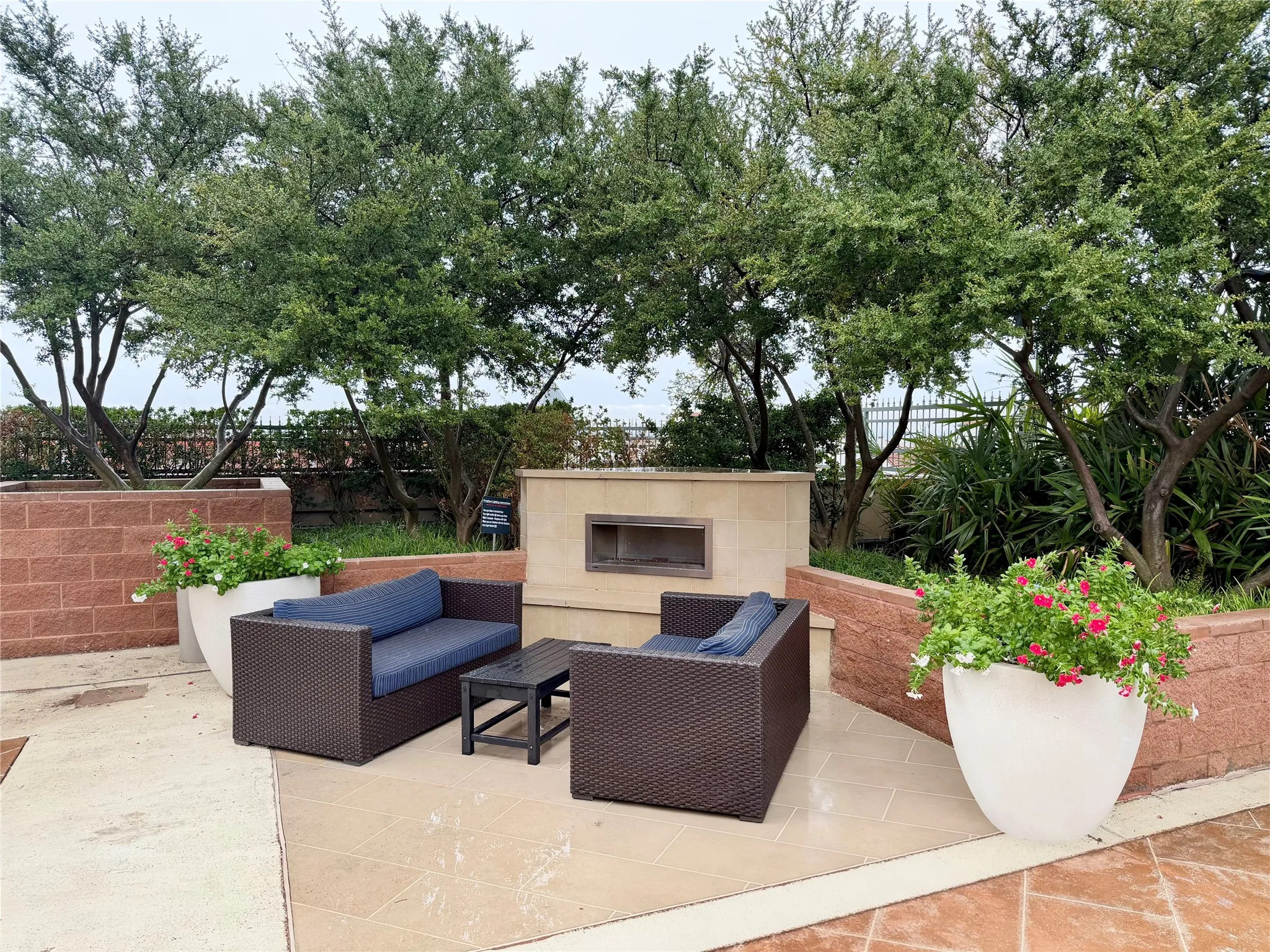 Tiled patio area with a built-in outdoor fireplace, surrounded by raised brick planters and mature trees