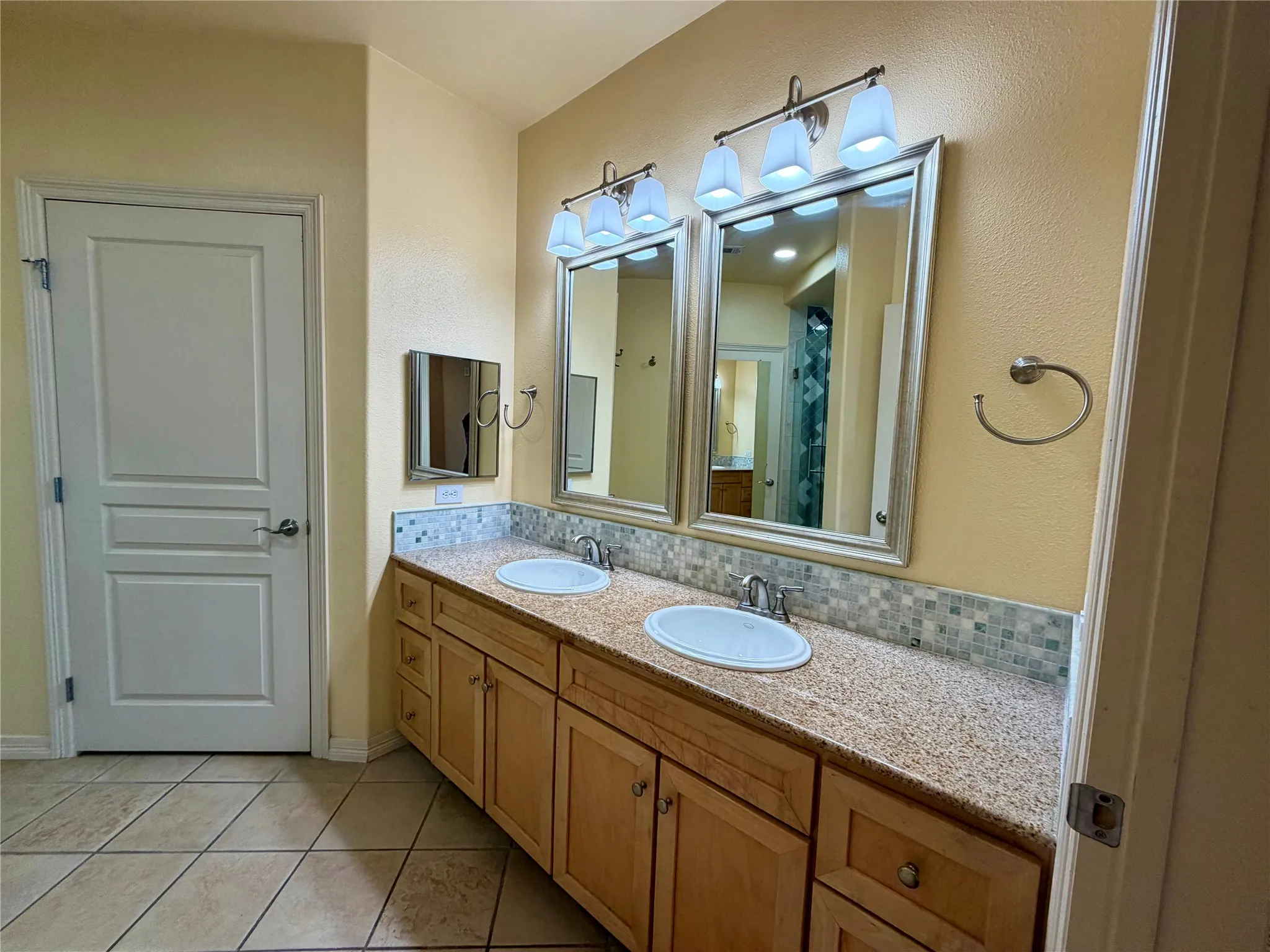Double vanity with integrated sinks, mosaic tile backsplash, light wood-finish cabinetry, and tiled flooring