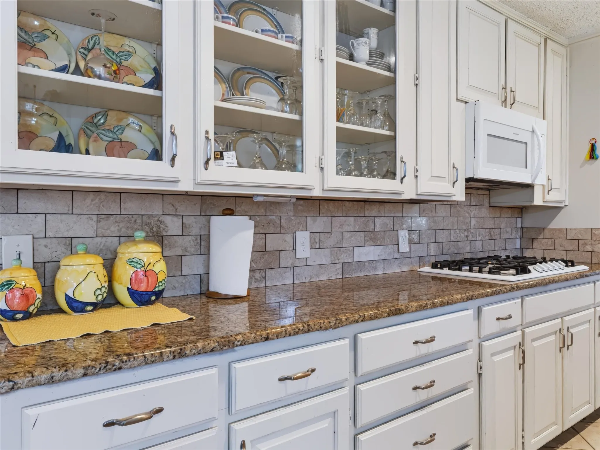 White cabinetry with glass-front upper doors, granite countertops, a tiled backsplash, built-in microwave, and a gas cooktop