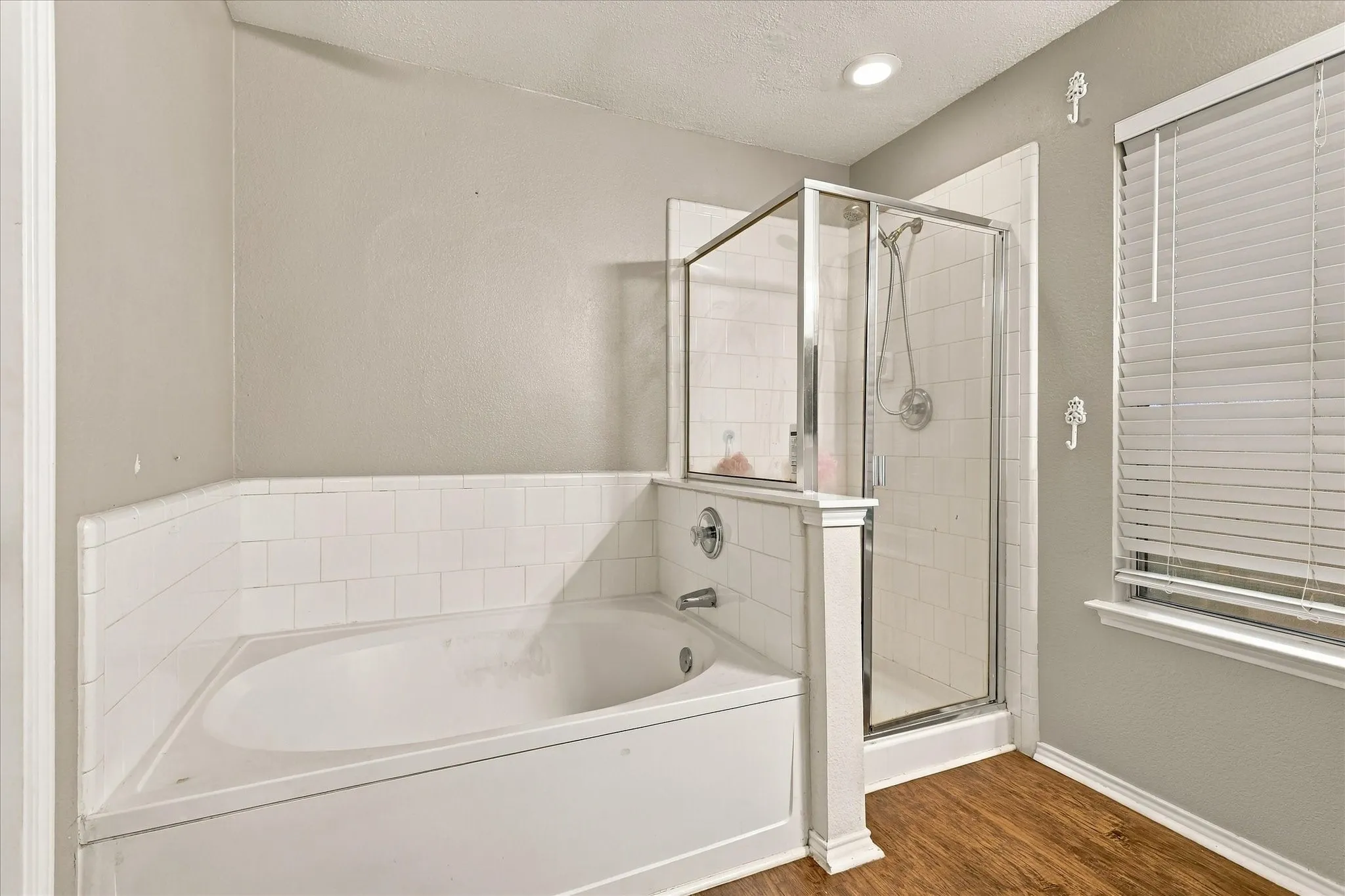 Built-in soaking tub with white subway tile surround, adjacent glass-enclosed shower, and wood-finish flooring