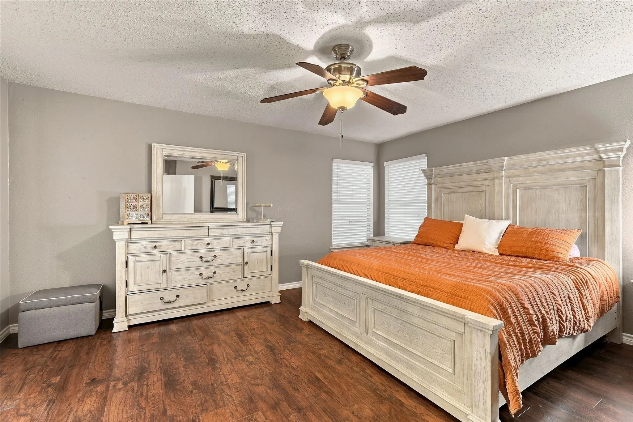 Spacious primary bedroom showcasing a white painted wood bed frame, wood-finish flooring throughout, and abundant natural light from corner windows with plantation shutters