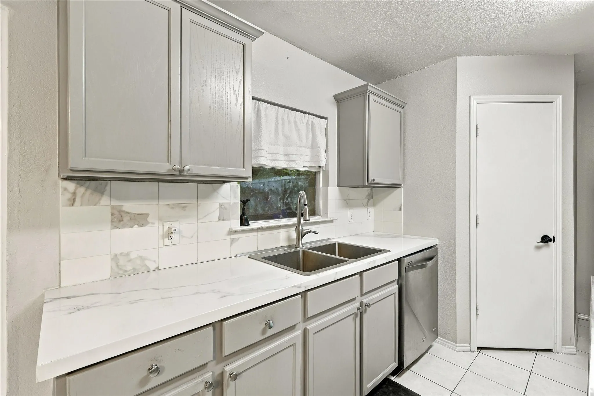 Gray shaker-style cabinetry with chrome hardware, dual basin stainless steel sink, stainless steel dishwasher, white countertops, and white tile flooring