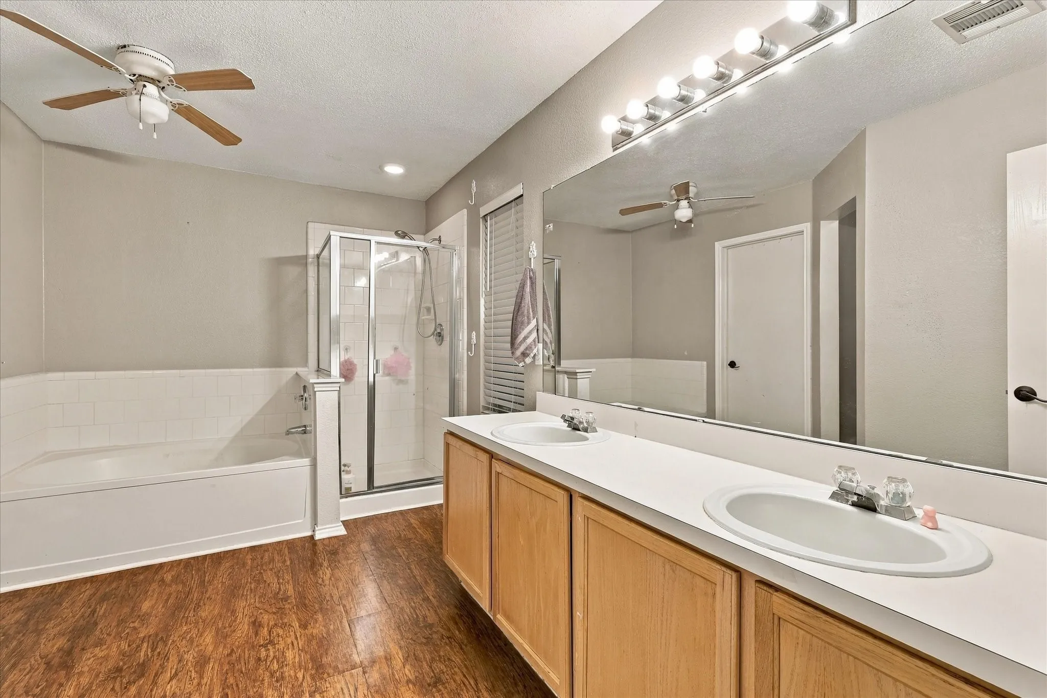 Double vanity with integrated sinks, large wall-mounted mirror, walk-in shower with glass enclosure, separate soaking tub with tile surround, and wood-finish flooring