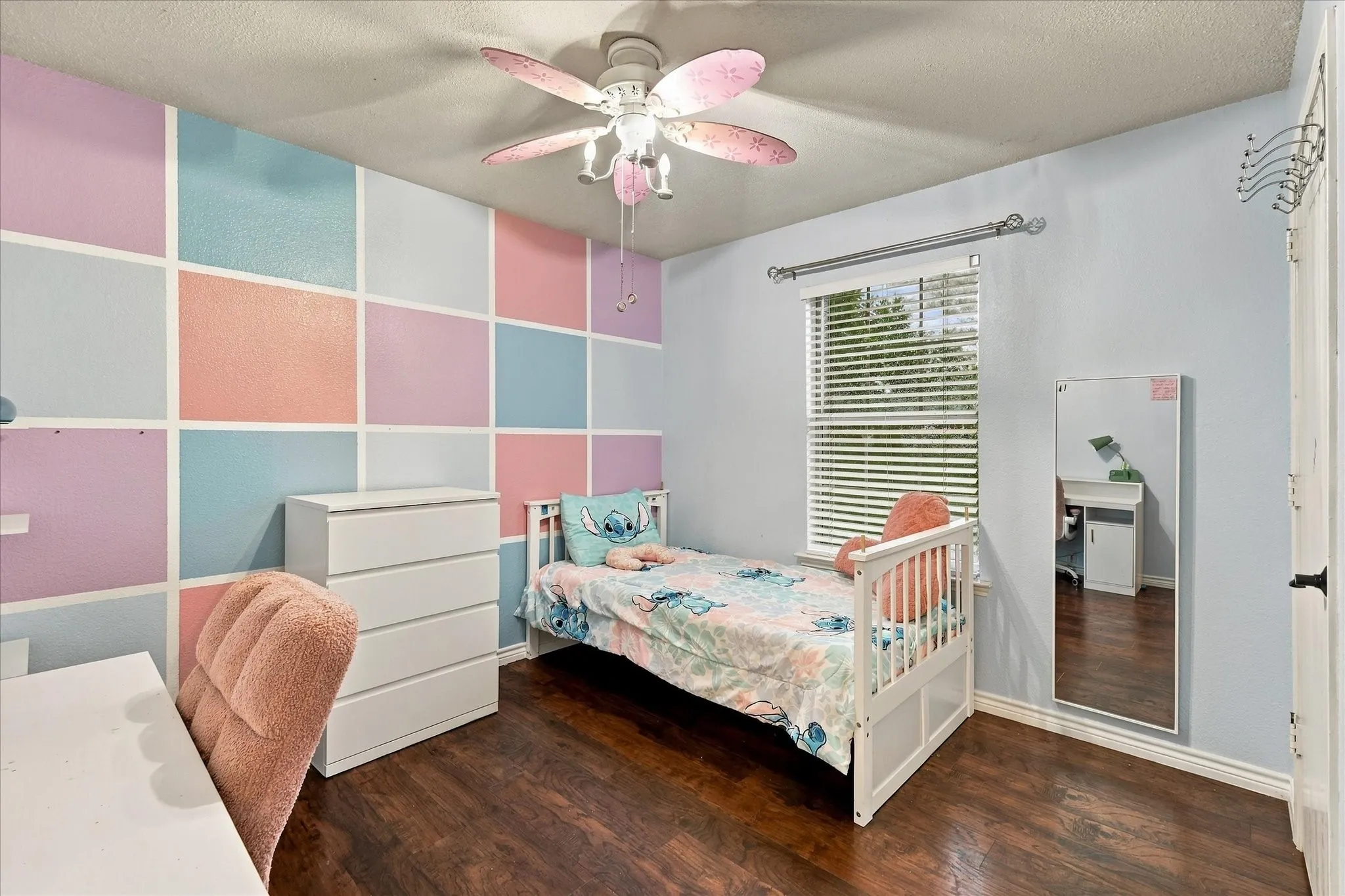 Wood-finish flooring, accent wall with a geometric pattern, ceiling fan with light fixture, window with blinds, and light-toned wall paint