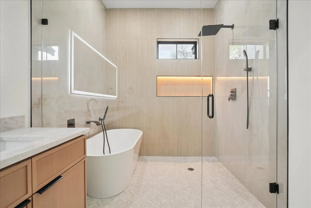 Freestanding soaking tub with floor-mounted faucet, illuminated vanity mirror, glass-enclosed shower with rain shower head, and recessed shower niche with accent lighting