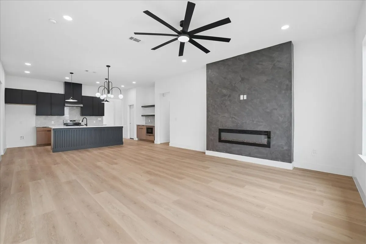 Open-concept living area with wood-finish flooring, a modern linear fireplace, and a sleek kitchen featuring dark cabinetry and a central island with a decorative slatted facade