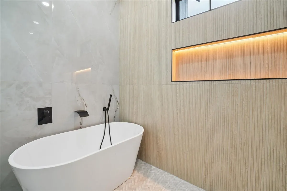 Freestanding soaking tub with matte black fixtures, full-height light-toned tile wall, and an illuminated recessed shelf