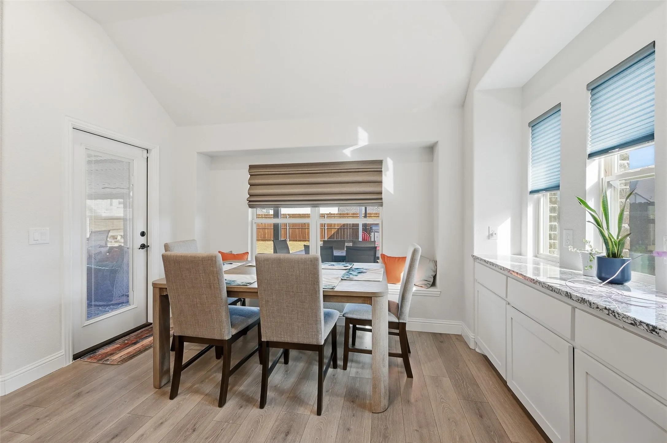 Dining area with vaulted ceiling, wood-finish flooring, built-in window seat, large window with shades, and white cabinetry with a stone-finish countertop