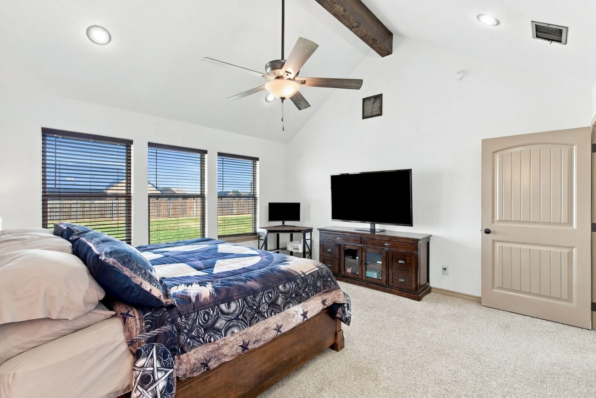 Vaulted ceiling with exposed wood beams, ceiling fan, recessed lighting, three windows with blinds, and light-colored carpeting