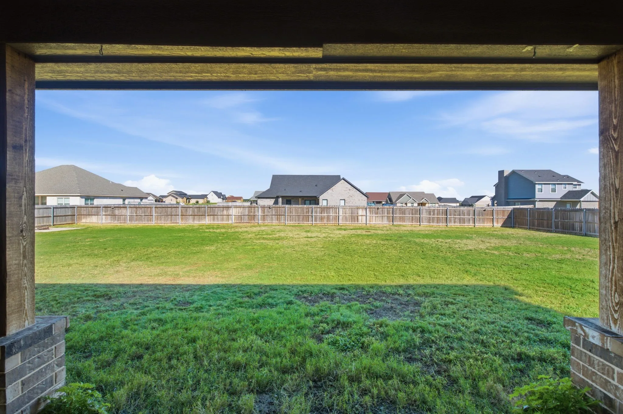 Covered patio with exposed wood beams and brick pilasters, opening to a large fenced yard with a wood privacy fence