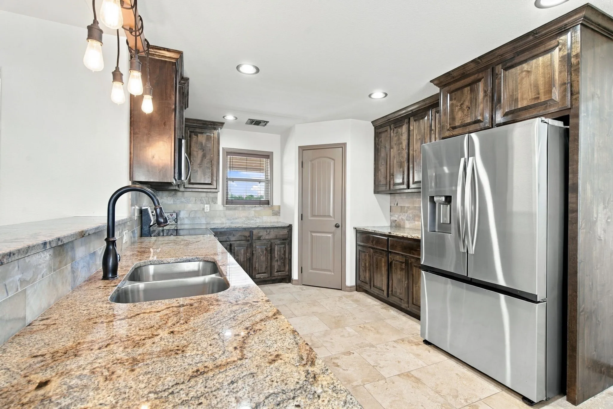 Kitchen featuring stone countertops, dark wood-finish cabinetry, a stainless steel refrigerator, a double basin sink with a black faucet, and recessed lighting