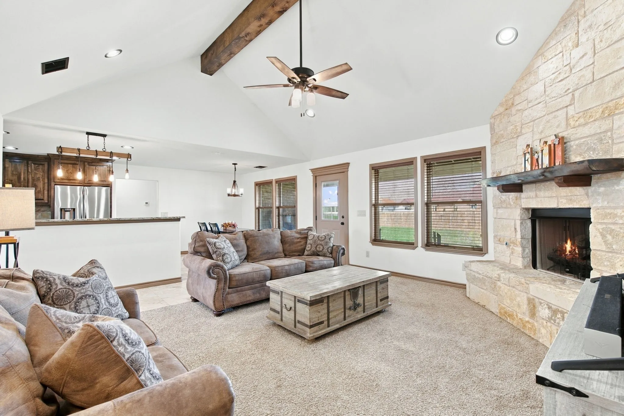 Vaulted ceiling with wood beam accent, stone-clad fireplace, built-in fireplace hearth, ceiling fan, and recessed lighting
