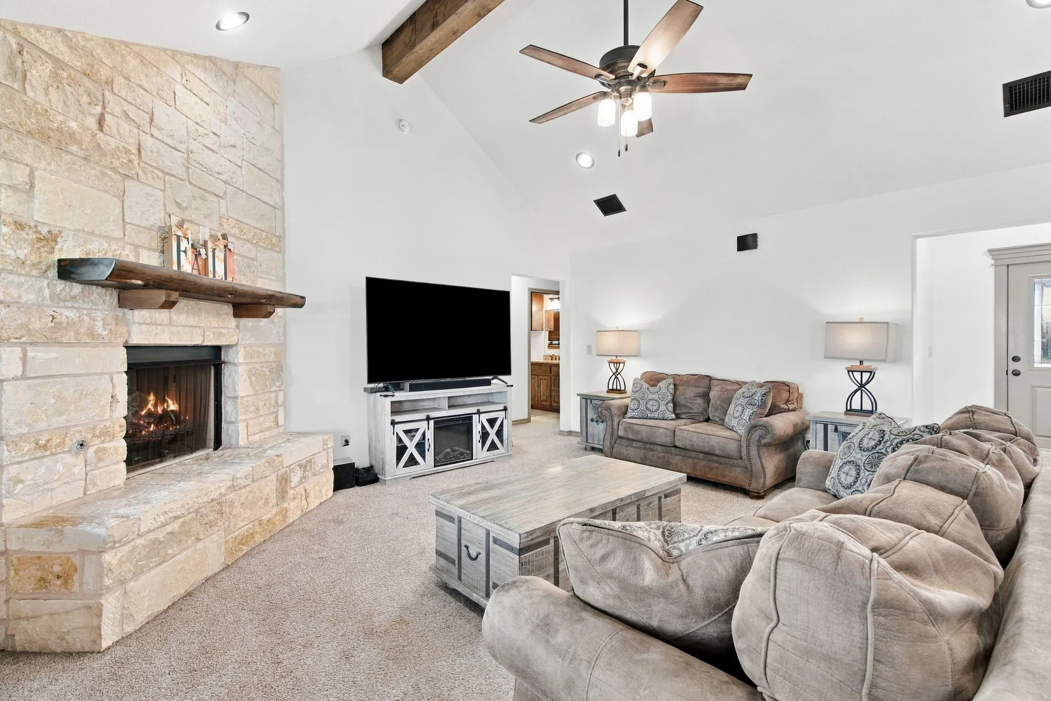 Stone-clad fireplace with a wood mantle, vaulted ceiling with exposed wood beams, ceiling fan with integrated lighting, and recessed lighting