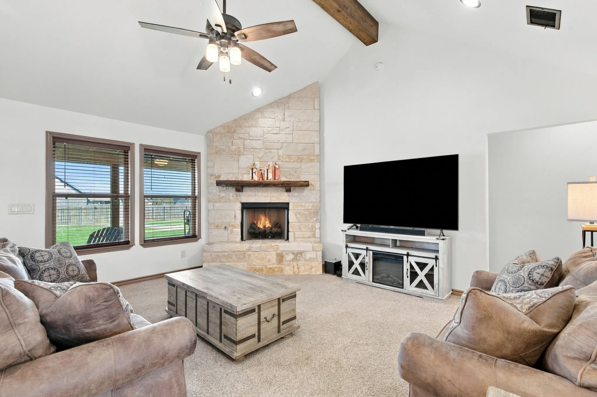 Vaulted ceiling with an exposed wood beam, stone-clad fireplace with a wood mantel, ceiling fan, and two windows with blinds