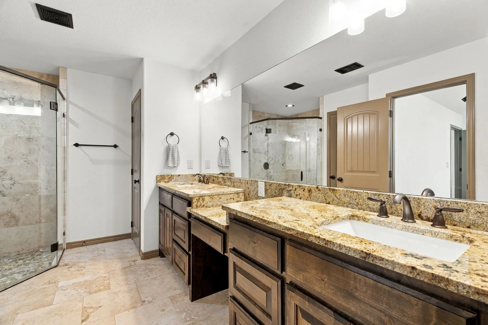 Dual vanity with stone countertops and undermount sinks, bronze finish plumbing fixtures, wood-finish cabinetry, and a walk-in shower with stone tile