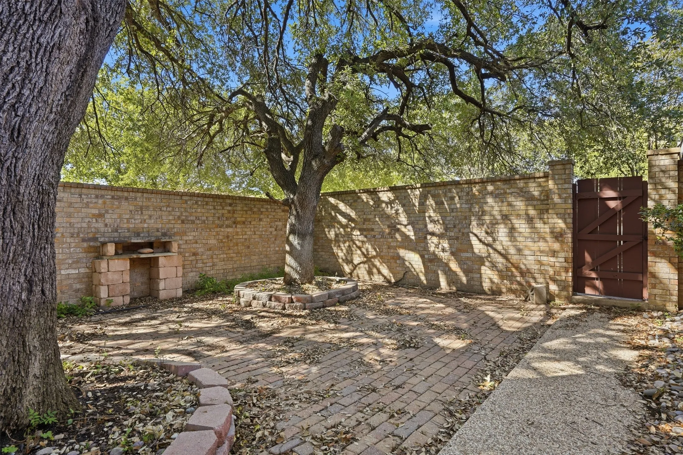 Enclosed brick-walled outdoor area featuring a brick paver pathway, mature trees, a built-in brick structure, and a wooden gate