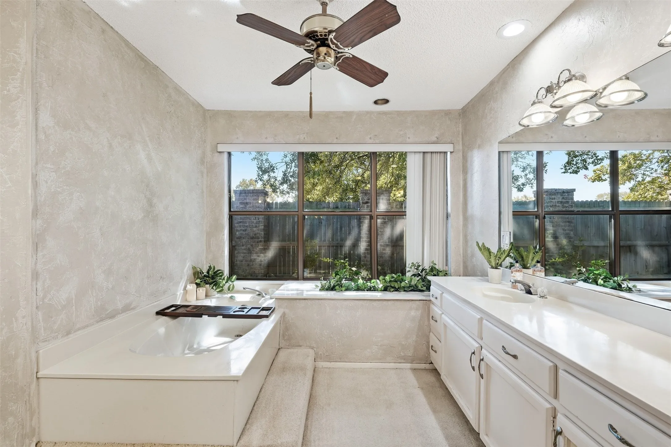 Dual-sink vanity with white cabinetry, built-in bathtub, ceiling fan, and large windows