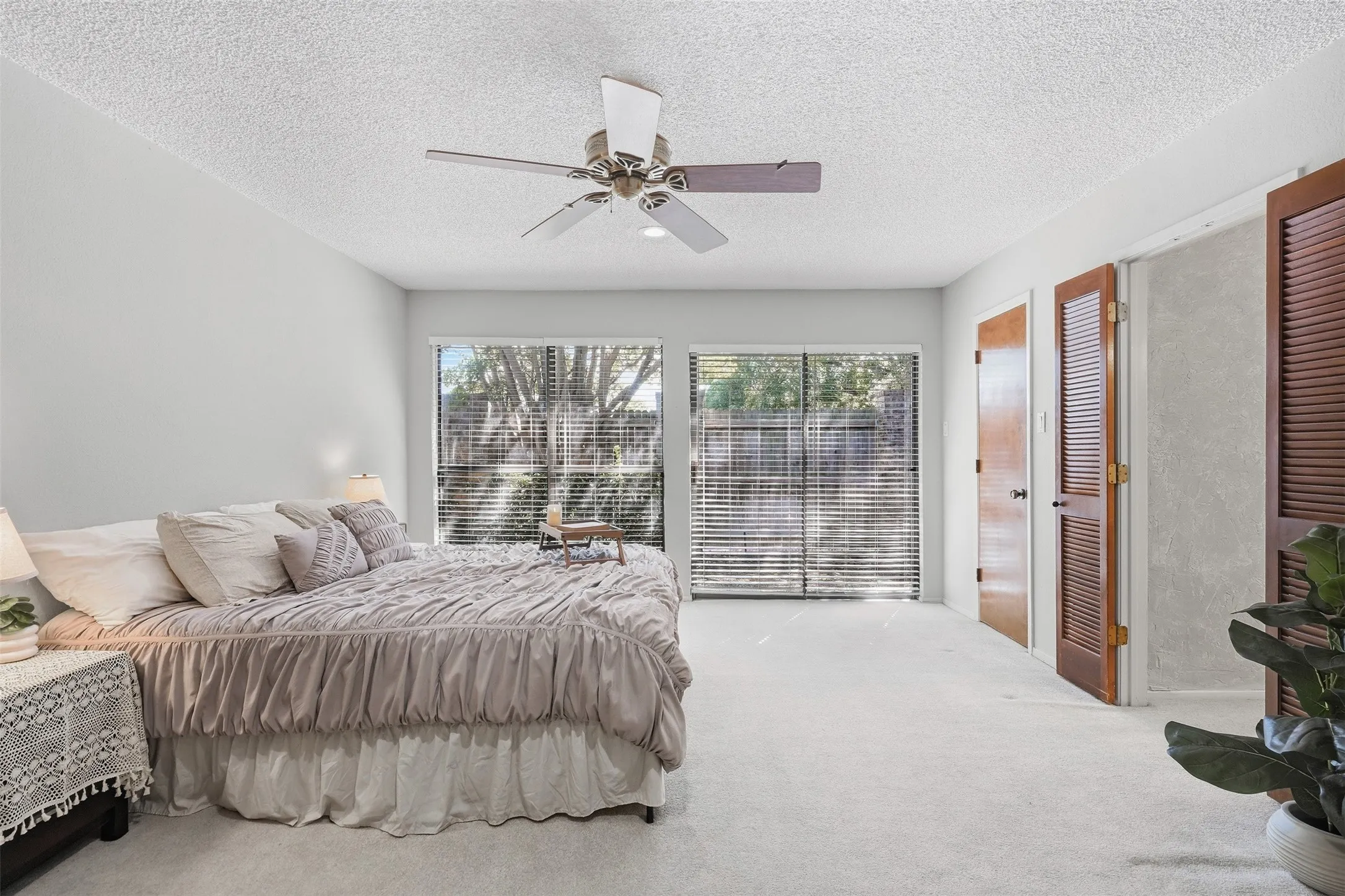 Dual sliding glass doors with horizontal blinds, ceiling fan with light fixture, light-toned carpeting, and wood-finish doors