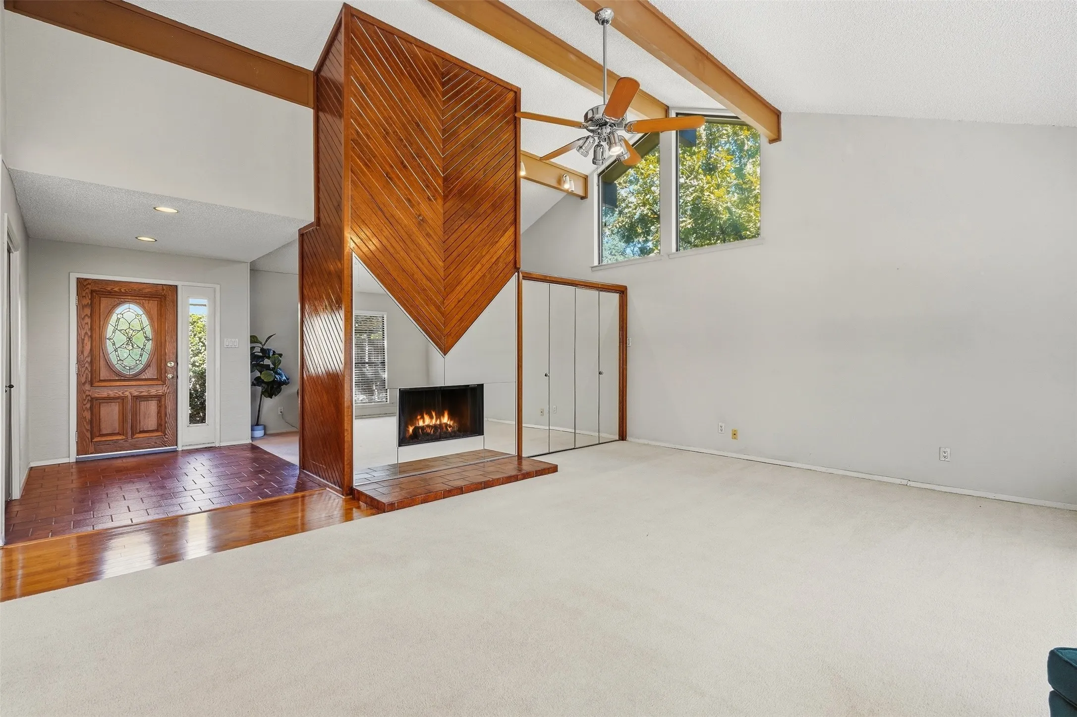 Vaulted ceiling with exposed wood beams, a prominent wood-paneled fireplace, large windows providing natural light, and a wood-paneled entry door with an oval glass insert