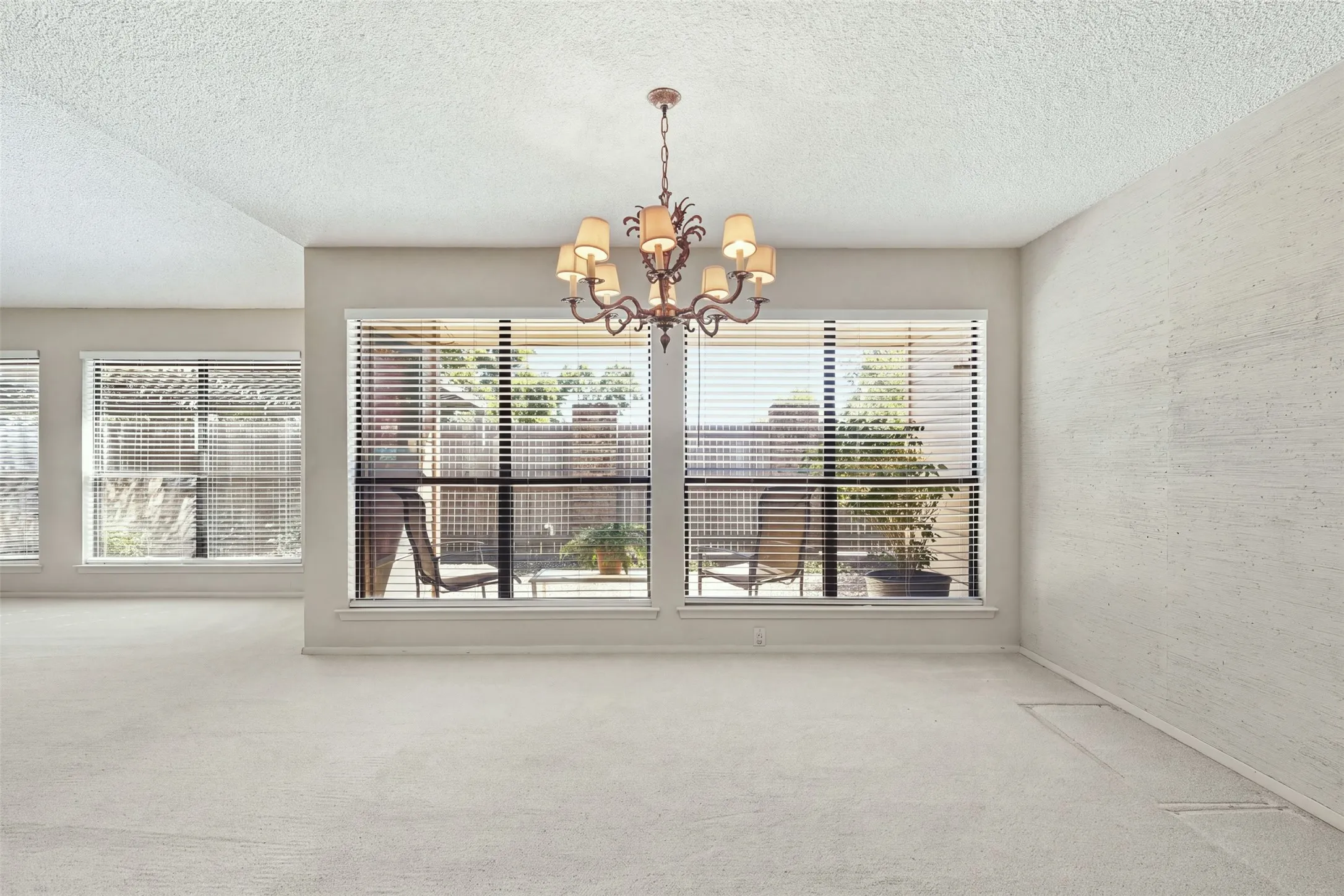 Vaulted ceiling, decorative chandelier, light-colored carpeting, multiple windows with horizontal blinds, and a private outdoor space