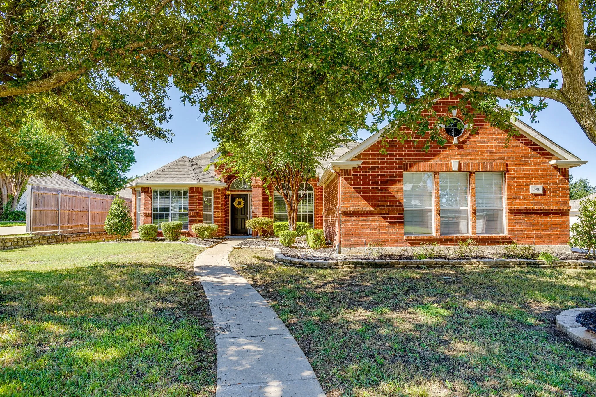 Brick facade residence with a gabled roofline, mature trees, a concrete pathway, and curated landscaping