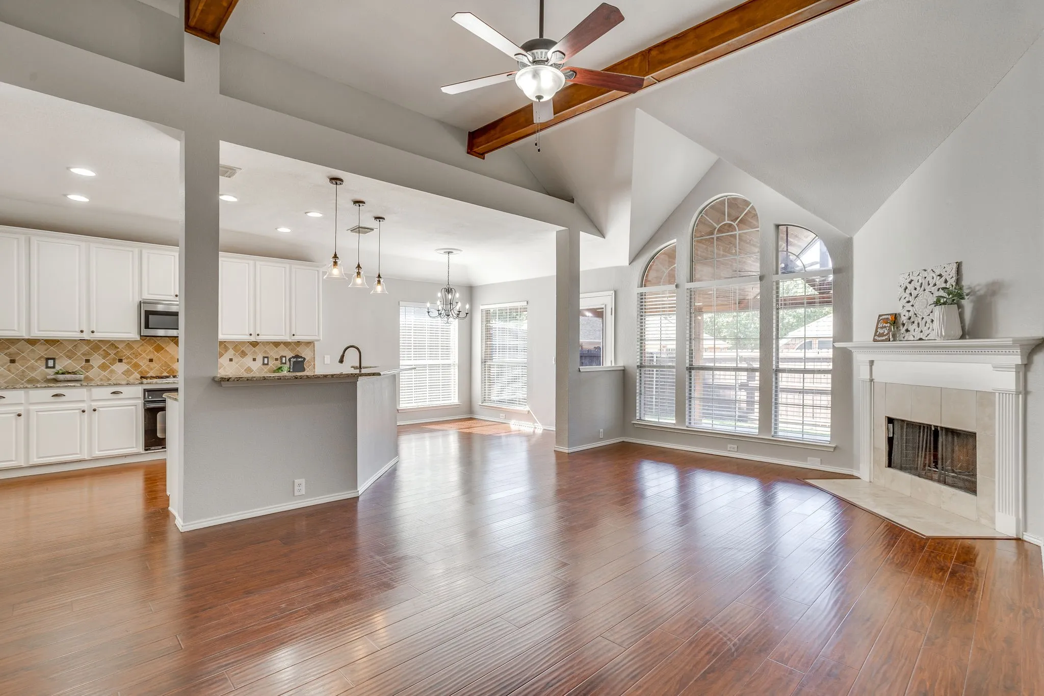 Vaulted ceiling with wood beams, wood-finish flooring, white kitchen cabinetry, a fireplace with a mantel, and arched windows